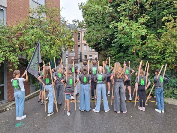 Les élèves de 6e, vus de dos, avec leurs échasses et le drapeau de l'école avant la Joute des enfants